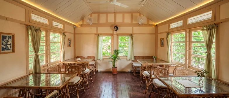 Wide view of the upstairs dining hall, with cane tables and leaded-glass windows opening to the trees